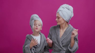 Mor stüdyo arka planına izole edilmiş kozmetik ürünler uygulayan çekici anne ve kızı otel bornozları poz veriyor. Reklamcılık.