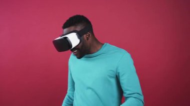 Naneli süveter giyen yakışıklı Afrikalı adam sanal gerçeklik gözlüğü takıyor, etrafa bakıyor. Genç, yakışıklı bir adam aşağı bakıyor, yukarı bakıyor, gülümsüyor. Görsel efektler. Pembe duvar.