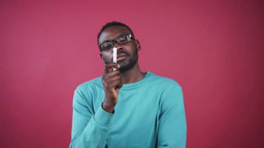 Siyah gözlüklü çekici Afrikalı genç bir adam kameraya bakıyor, elinde kalemle. Pembe duvarın arka planında kameraya bakan düşünceli bir adam..