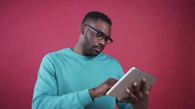 Çekici Afrikalı adam çalışıyor, dijital tablet kullanıyor, doğrudan cihazın ekranına bakıyor. Siyah gözlüklü, kameraya bakan sempatik genç bir adam. Pembe duvar.