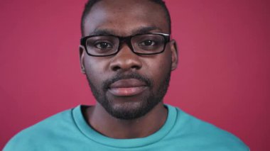 Gözlüklü çekici Afrikalı genç adam kameraya dikkatlice bakıyor, gülümsüyor. Pembe duvarın arka planında kahverengi gözlü hoş bir adam. Yakın plan..