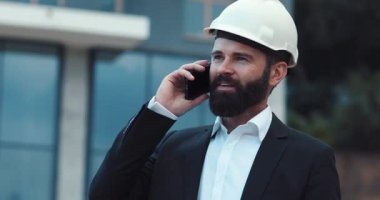 Kasketli bir iş adamı inşaat arka planında telefonla konuşuyor. Meslek konsepti, modern iletişim araçları. İş yapıyorum. Erkek portresi