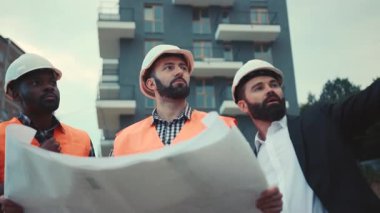 Miğferli inşaat işçileri ve işadamları yapılan işi tartışıyor, proje planıyla kontrol ediyorlar. Meslek konsepti, aktif tartışma. Takım çalışması..