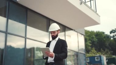 Takım elbiseli ve kasklı ciddi sakallı bir işadamı tabletini kullanarak inşaat alanında yürüyor. Profesyonel, dijital projeksiyon, modern teknolojiler. Erkek portresi