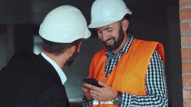 Kasklı yakışıklı adam inşaat alanındayken aktif olarak akıllı telefon kullanıyor, bir iş adamı ona geliyor, şakalar yapıyor, sırtını tokatlıyor. Ortak çalışma.