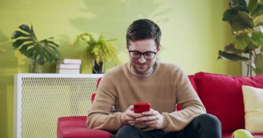 Genç bir adam, bitkilerle ve modern dekorlarla dolu rahat bir oturma odasında rahatlarken akıllı telefonunun keyfini çıkartıyor.