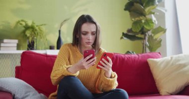 Stresli genç bir kadın, iki akıllı telefondan evde tartışırken mesajlaşıyor. Yakın planda, kafası karışık beyaz kız umutsuzluk içinde kanepede oturuyor..