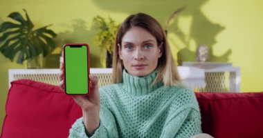 Bir kadın elinde parlak yeşil ekranlı akıllı bir telefon tutuyor. Etrafında modern ve modaya uygun bir yaşam alanı sunuyor.
