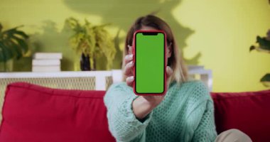 Sarışın kadın evinde elinde sahte akıllı telefon, yeşil ekran e-ticaret web sitesi, rahat içinde dikey pozisyonda. Sıradan insanlar ve teknoloji..
