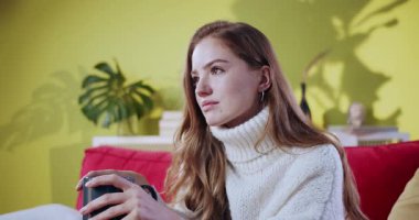 Güzel, kızıl saçlı, elinde çay fincanı tutan genç bir kadın evinde mutlu bir şekilde gülümsüyor. Neşeli kızıl kız, sıcak içecekle ısınıyor, rahat bir şekilde dinleniyor..