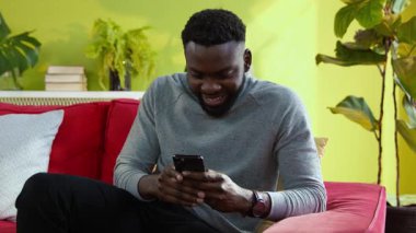 Modern akıllı telefondan finans haberleri okuyan mutlu Afrikalı adam ev oturma odasında zafer servetini kutlarken aşırı sevinçten havalara uçuyor..