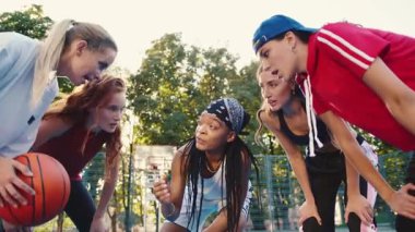 Bir grup genç kadın bir basketbol sahasında birlikte strateji oluşturuyorlar, elinde sıkı bir topla.