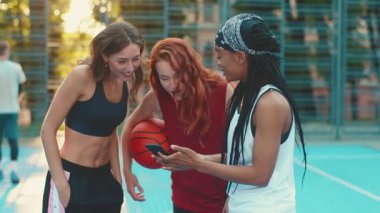 Üç arkadaş aktif olarak nişanlanmışlar ve telefonlarını kontrol ederken neşeli bir basketbol sahasında eğleniyorlar.