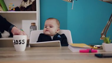Şık bir takım elbise giymiş sevimli bir bebek, parlak, renkli bir dekorla çevrili bir tabletle bir masada oturuyor.