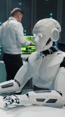 Sunucu odasında insanlarla çalışan masaüstü bilgisayar programlama yazılımı kullanan sibernetik beyaz robot. Veri merkezi. Yapay zeka.