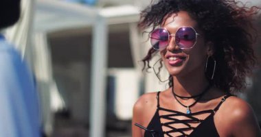 Çekici, genç, güzel bir kadın. Güneş gözlüklü bir adamla konuşuyor. Gülümsüyor, gülüyor, rahatlıyor.