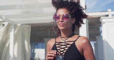 Güneş gözlüklü çekici genç bir kadın gülümsüyor gülüyor sahildeki bir partide kokteyl içtiği için çok ırklı gün batımını rahatlatıyor.