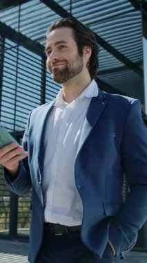 Mavi ceketli, iş merkezinin yakınlarında yürüyen mutlu ve zeki bir genç adam görüntüsü. Yakışıklı, sakallı, beyaz iş adamı elinde akıllı bir telefonla ekrana bakıyor ve gülümsüyor. Başarı. Açık hava.