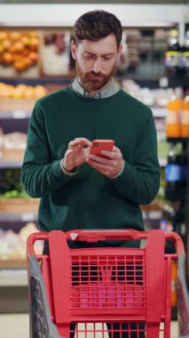 Genç beyaz sakallı adam müşterisi cep telefonu uygulamasını kullanarak süpermarkette alışveriş arabasıyla bekliyor. Alışverişe. Kontrol listesi. Market..