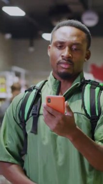 Kafası karışmış kurye Afro-Amerikan bir adam dcutomer siparişiyle ilgili bir yanlış anlaşılma yaşıyor süpermarkette akıllı telefon kullanarak omuz silkiyor.,