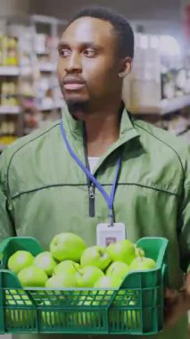 Afro-Amerikan işçi süpermarketinin portresi kameraya gülümsüyor, elinde bir sepet dolusu elmayla markette çalışırken mutlu görünüyor..