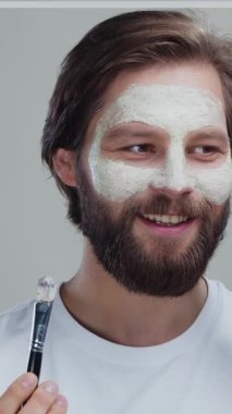 Beyaz kilden maske takan neşeli bir adamın portresi kozmetik fırçayla gri arka planda izole edilmiş akıllı telefondan kız arkadaşıyla konuşuyor..