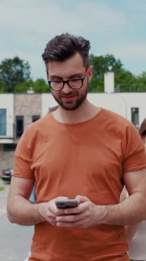 Turuncu gömlek giyen genç bir adam dışarıda rahat bir ortamda dinlenirken akıllı telefonuyla meşgul oluyor.