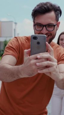Şık gözlükler takan neşeli genç bir adam parlak, güneşli bir ortamda akıllı telefonuyla mutlu bir selfie çekiyor.