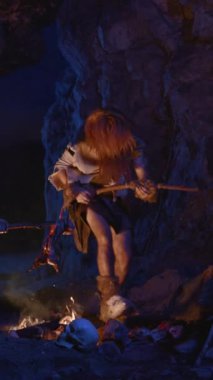 Elinde silah tutan bir mağara adamı karanlık mağara çevreleriyle çevrili titreyen bir ateşin yanında cesurca duruyor.