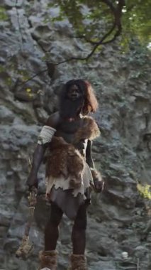 İlkel giysiler içinde, kayalık bir arazinin ortasında cesurca duran vahşi bir savaşçının çarpıcı tasviri.