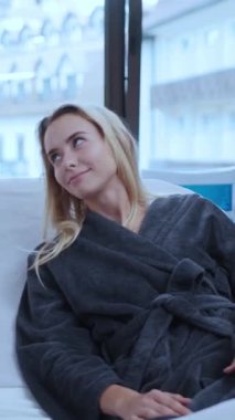 Huzurlu bir an, rahat bir odada, yumuşak bir bornozla pencereden dışarı dikkatlice bakan bir insanın görüntüsü.