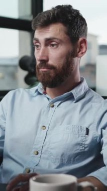 Bakımlı sakallı genç bir adam parlak, modern bir kafe ortamında kahvesini yudumlamaktan zevk alır.