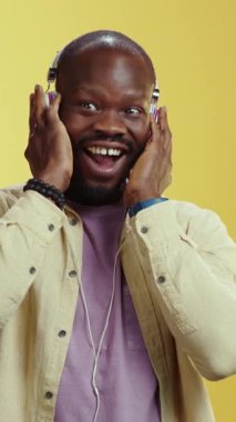 Bir adam rahat bir şekilde oturur, kulaklıkla eğlenceli müziğe dalmış, canlı sarı bir arka plana kurulmuştur.