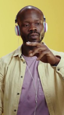 Müzik dinleyen, şık kulaklıklar takan, kendine güvenen bir adam, canlı sarı bir arkaplan karşısında duruyor..