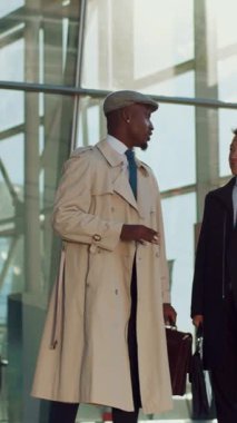 Şık bir işadamı, şehir merkezindeki cam bir binanın yanında trençkot ve kep giyerken özgüvenle el kol hareketi yapar.