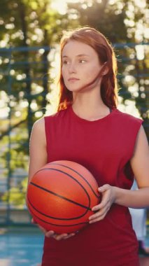 Odaklanmış genç bir kız elinde bir basketbol topuyla güvenle dışarıda duruyor, kararlılık ve hırs saçıyor.