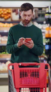Bir adam manavın koridorlarında alışveriş yaparken akıllı telefonunu kontrol eder ve eşyaları inceler.