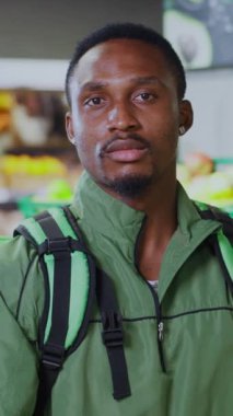 Genç bir adam markette duruyor, yeşil bir ceket giymiş, ve yanında bir sırt çantası taşıyor.