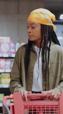 Düşünceli bir müşteri, canlı ve renkli bir market koridorunda elinde alışveriş arabasıyla düşünceli bir şekilde durur.