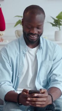 Neşeli genç bir adam, rahat ve rahat bir ev ortamında, akıllı telefonuyla mutlu mesut meşgul olur.