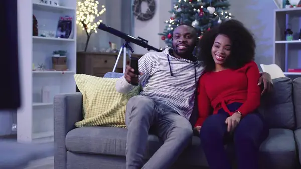 Mutlu Afrikalı çift akşamları dinleniyor. Gençler kanepede oturur ve dekore edilmiş Noel ağacının arka planında televizyon izlerler..