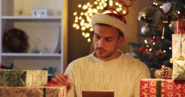 Sakallı, Noel Baba şapkası takan, neşeli bir geçmişi olan yakışıklı bir genç. Hediyeleri ve tablete dokunmayı düşünen adam. Gülümsüyor.