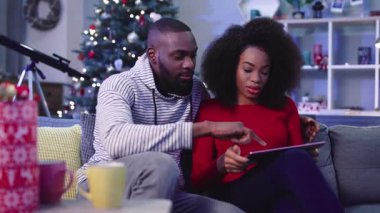 Birbirine aşık çift tablet gösterisinde birbirine dokunuyor. Gençler yeni yıl ağacının arka planında koltukta oturmuş süslenmişler. Çıkma.