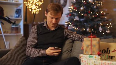 Kız arkadaşıyla mesajlaşan yakışıklı, beyaz bir adam. Çelik yelek giymiş genç çekici bir adam arka planda oyuncaklar ve toplarla harika bir Noel ağacı hakkında görüntülü sohbet ediyor..