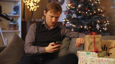 Bir sürü güzel kutuyla kanepede oturan yakışıklı beyaz adam. Yelek giymiş genç bir adam, görüntülü konuşma yapıyor. Adam eğleniyor. Kutlama atmosferi.