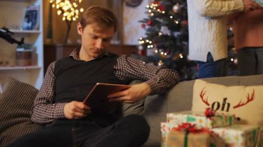 Genç ve çekici bir adam dijital tablet ekranına bakıyor ve güzel kutuları düşünüyor. Aşık genç bir çift. Noel atmosferi.