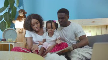 Afrika kökenli aile, oturma odasında yatakta oturup hafta sonunu evde birlikte geçiriyorlar. Heyecanlı ebeveyn ve kızın birlikte güzel bir kitap okuduğu portre..