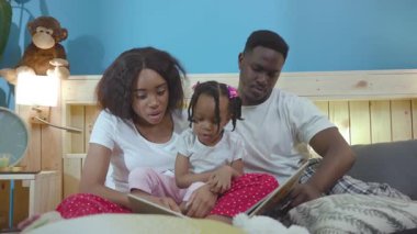 Siyahi Afrikalı sevecen ebeveynler yatakta uzanmış heyecanlı hikayeler okuyan bir çocuk yetiştirdiler. Güzel bir aile portresi..