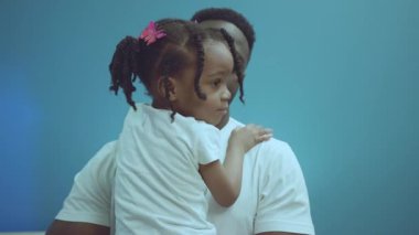 Neşeli bir Afrikalı Amerikalı babanın portresi. Mavi arka planda izole edilmiş sevimli küçük kızı kucağına alıp kucaklıyor..