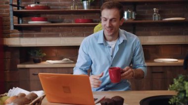 Mutfaktaki dizüstü bilgisayardan olumlu haberler okuyan başarılı girişimci gülümsüyor. Masada oturmuş, ekrana bakıp kahve içen gülümseyen genç adamın portresi..